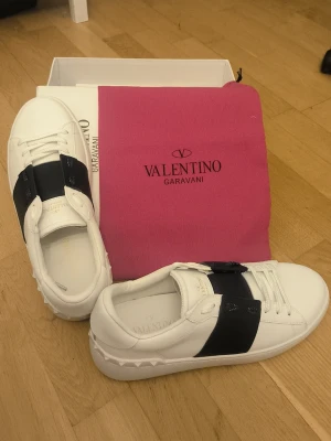 Valentino Garavani vita sneakers - Säljer ett par vita sneakers från Valentino Garavani med bred marinblåa läderdetalj över snörningen och subtila nitar på hälen. Skorna har en klassisk låg profil och är tillverkade i slätt läder. Perfekta för dig som gillar exklusiv och stilren design.