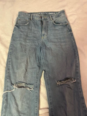 Ljusa jeans från Bikbok - Säljer ett par ljusblå jeans från Bikbok med hög midja och raka ben. Jeansen har hål på båda knäna för en trendig look. Klassisk femficksmodell och perfekta för dig som gillar avslappnad streetstil.