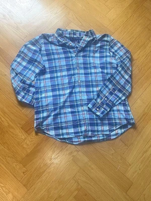 Blårutig skjorta med långa ärmar - Säljer en klassisk blårutig skjorta med långa ärmar och knäppning framtill. Skjortan har en button-down krage och manschetter med knappar. Mönstret består av olika nyanser av blått, vitt och lite rött. Perfekt för en avslappnad men ändå stilren look.