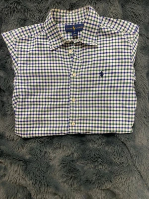 Rutig skjorta från Ralph Lauren - Klassisk rutig skjorta från Ralph Lauren i blått, grönt och vitt. Skjortan har button-down krage, broderad logga på bröstet och vita knappar framtill. Tillverkad i bomull för en skön och luftig känsla. Perfekt för en stilren och avslappnad look. Storlek: 12