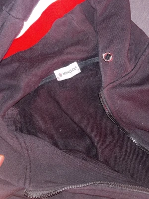 Svart Moncler hoodie med dragkedja - Säljer en svart hoodie från Moncler med dragkedja framtill och huva. Huvan har snygga röda och vita ränder som ger en sportig vibe. Tillverkad i mjukt bomullstyg och har ribbade muddar vid ärmslut och nederkant. Perfekt för dig som gillar stilrena och exklusiva plagg.