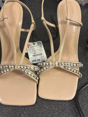Beige pumps med strass från Zara - Säljer ett par snygga beige pumps från Zara med tunna remmar och fyrkantig tå. Skorna har dekorativa strassdetaljer framtill som ger extra bling. Tillverkade i konstläder med smal klack och elegant design. Perfekta för dig som vill sticka ut.