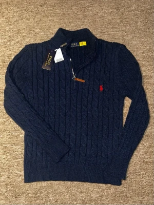 Mörkblå stickad tröja Polo Ralph Lauren - Kabelstickad mörkblå tröja från Polo Ralph Lauren med halv dragkedja och röd broderad logga på bröstet. Tröjan har hög krage, långa ärmar och ribbade muddar. Perfekt för dig som gillar klassisk och stilren design.
