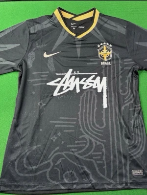 Svart Brasilien x Stüssy tröja - Unik svart kortärmad tröja från Nike i samarbete med Stüssy och Brasilien. Tröjan har gul detalj vid kragen, Brasilien-emblem på bröstet och Stüssy-logga i vitt framtill. Tillverkad i lätt, andningsbart material perfekt för sport.