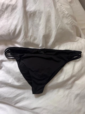 Svarta bikiniunderdel med sidosnörning - Säljer en svart bikiniunderd från Tezenis med trendiga dubbla snören i sidorna i storlek S. Klassisk modell med låg midja och enkel, stilren look. Perfekt för stranden eller poolen.