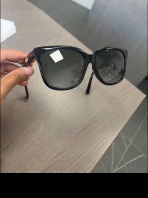Svarta solglasögon från Gucci - Snygga svarta solglasögon från Gucci med klassisk cat eye-form och mörka glas. Bågarna är i blank svart plast och har en liten gulddetalj vid gångjärnet. Perfekta för att höja din stil på sommaren.