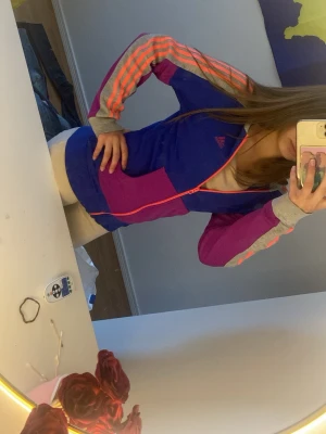 Färgglad Adidas zip hoodie - Säljer en sportig hoodie från Adidas i blått, lila och rosa med dragkedja framtill. Tröjan har meshfoder, två fickor och klassiska Adidas-ränder i neonrosa på ärmarna. Perfekt för träning eller chill. Märke synligt på bröstet.