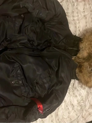 Svart bomberjacka Alpha Industries - Säljer en svart bomberjacka från Alpha Industries med klassisk röd 'Remove Before Flight'-tag på ärmen. Jackan har flera fickor, dragkedja och knappar samt en huva med fuskpäls. Materialet är glansigt och slitstarkt, perfekt för en cool streetstil. Trasig vid kedjan går dock att fixa enkelt 