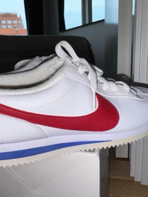 Nike Cortez vita och röda sneakers - Klassiska Nike Cortez sneakers i vitt skinn med röd swoosh och blå detalj på sulan. Lågt skaft, vita skosnören och sula med sågtandad kant. Perfekta för en clean och retro look.