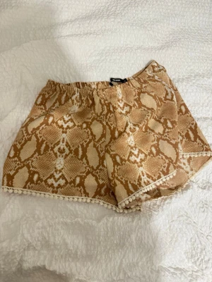 Beige shorts med ormmönster från Bik Bok - Snygga beige shorts från Bik Bok med trendigt ormmönster och dekorativ spetskant längst ner. Perfekta för varma dagar och ger en cool vibe till din outfit. Resår i midjan för skön passform.