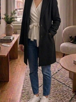 Svart klassisk ullrock - Stilren svart rock med rak passform och två knappar framtill. Rocken har diskreta fickor på sidorna och en klassisk krage. Perfekt för dig som vill ha en tidlös och enkel look. Materialet ser ut att vara ullblandning och modellen är knälång.