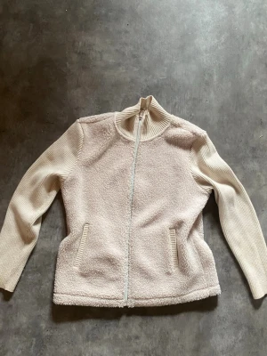Väst med armar - Säljer en beige väst/jacka med dragkedja framtill och två fickor. Jackan har mjuk, fluffig teddy på kroppen och ribbstickade ärmar samt krage för extra stilpoäng. Perfekt för dig som vill ha en mysig och trendig look.