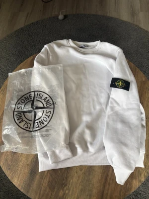 Vit sweatshirt från Stone Island - Vit sweatshirt från Stone Island med klassisk rund hals och ikonisk logotyp-patch på vänster ärm. Tröjan har ribbade muddar vid ärmslut och nederkant. Perfekt för en clean och stilren look. Bra kvalité för att va ifrån 🐼