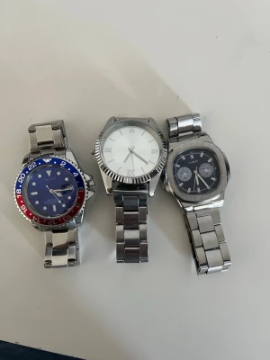 3 st herrklockor i silverfärg - Säljer tre stilrena herrklockor med metallarmband. En har blå urtavla med röd och blå vridring, en har klassisk silverfärgad urtavla med romerska siffror och en har svart urtavla med kronografdetaljer. Perfekta accessoarer för dig som gillar snygga klockor. Den silvriga är helt oanvänd men dom andra är använd skriv om du är intresserad eller har några frågor 