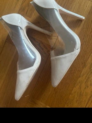 Vita pumps med transparent detalj - Säljer ett par vita pumps från Nly shoes med spetsig tå och transparenta sidor i plast. Klackarna är smala och eleganta, vilket ger en modern och trendig look. Perfekta för dig som vill sticka ut med en unik design.