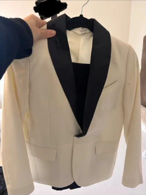 Vit kostym med svarta byxor från H&M - Snygg vit kavaj från H&M med kontrasterande svarta slag och klassisk passform. Kavajen har fickor framtill och en bröstficka, perfekt för en stilren look. Har en elegant design som sticker ut. Vanlig svarta kostymbyxor inkluderad.