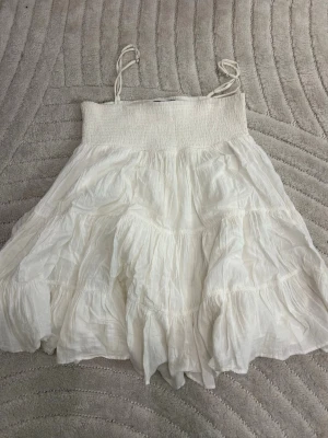 Vit sommarklänning från Zara - Säljer en söt vit kortklänning från Zara med tunna axelband och smock upptill. Klänningen har volangig kjol och är gjord i ett lätt, luftigt bomullstyg som passar perfekt till varma dagar. Enkel och stilren design som är lätt att matcha.