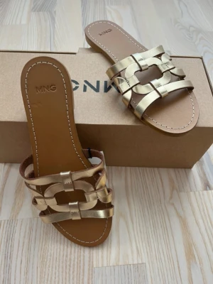 Guld lädersandaler  från Mango, 36 - Snygga sandaler från Mango i metallic guld med flätad design över foten. Öppen tå och platt sula i brunt konstläder. Perfekta för sommaren och ger en trendig touch till din outfit.