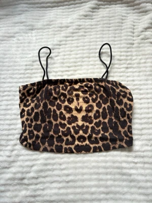 Leopardmönstrad bandeau topp - Säljer en leopardmönstrad bandeau topp med tunna axelband. Toppen har en croppad passform och är perfekt för dig som vill sticka ut. Materialet känns mjukt och stretchigt, troligen polyester eller syntet. Supersnygg att styla till jeans eller kjol.