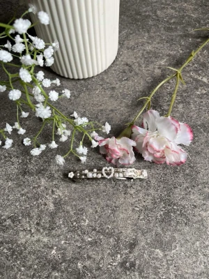 Silverfärgat hårspänne med pärlor - Säljer ett stilrent hårspänne i silverfärg dekorerat med små vita pärlor och ett hjärta i mitten. Perfekt accessoar för att piffa till frisyren och ge en söt touch till din look. Dessa spännen är handgjorda