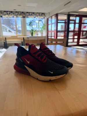 Nike Air Max 270 svart/röd sneakers - Nike Air Max 270 sneakers i svart och rött med vit sula och synlig Air-enhet i hälen. Ovandelen är i mesh för bra ventilation och har tydliga Nike-loggor på sidan och tungan. Snygg och sportig design med lågt skaft och röda detaljer.