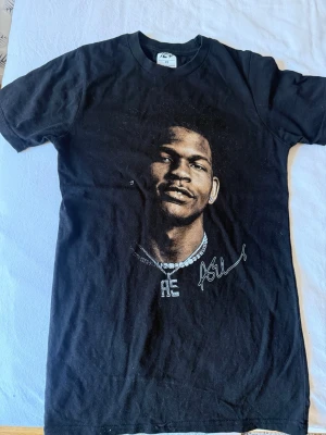 Svart Anthony Edwards t-shirt med tryck och signatur - Säljer en svart t-shirt med coolt tryck av en person på framsidan och en vit signaturdetalj. T-shirten har rund hals och korta ärmar, perfekt för en avslappnad streetwear-look. Materialet känns mjukt och skönt mot huden.