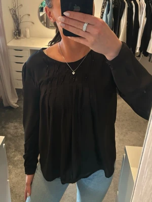Svart blus med veckad front - Stilren svart blus med rund halsringning och fina veckdetaljers framtill med små stenar. Blusen har långa ärmar och ett mjukt, lätt tyg som ger ett snyggt fall. Perfekt att matcha med jeans eller kjol för en trendig look.