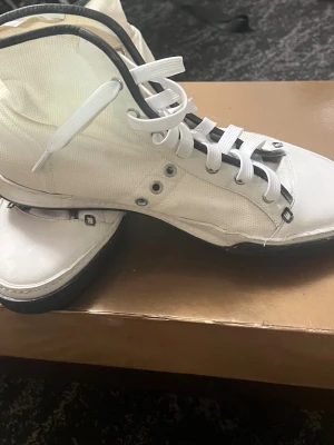 Vita high top sneakers med svart detalj - Säljer ett par vita high top sneakers med svarta detaljer och vita skosnören. Skorna har en canvasliknande ovandel, svart kant runt öppningen och svart sula. Märke syns på sidan av skon. Perfekta för en clean och stilren look.