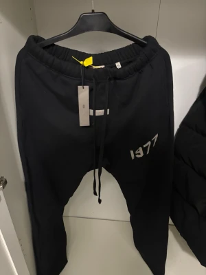 Svarta Fear of God Essentials mjukisbyxor - Säljer ett par svarta mjukisbyxor från Fear of God Essentials med trycket '1977' på vänstra benet. Byxorna har dragsko i midjan och ribbade muddar vid bensluten. Perfekta för en avslappnad och trendig look. Storkek Xl och L