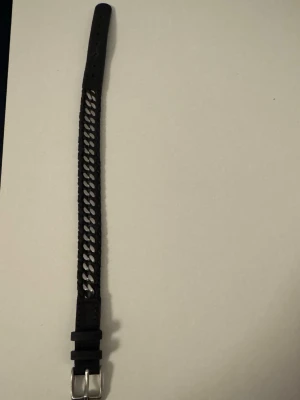 Svart flätat armband med silverdetaljer - Snyggt svart armbandmed flätad design och silverfärgat spänne. Armbandet har dekorativa hål längs hela längden och passar perfekt till en enkelt outfit. Materialet känns som skinn och ger en stilren look.