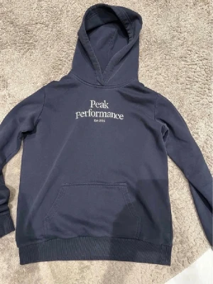 hoodie peak Performance - Snygg mörkblå hoodie från Peak Performance med vit logga och text på bröstet. Klassisk känguruficka framtill och ribbade muddar vid ärmslut och nederkant. Tillverkad i mjukt bomullsmaterial och har en stor huva för extra komfort. Strolek 170cm