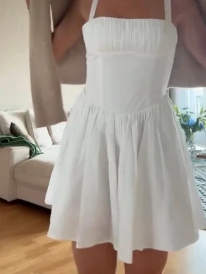 Zara klänning - Superfin vit kort klänning med smala axelband och rynkad överdel. Klänningen har en utställd kjol och är perfekt för varma dagar. Tillverkad i ett lätt och luftigt material som känns bekvämt mot huden.