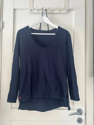 V-ringad tröja från Ralph Lauren - Mörkblå långärmad tröja från Ralph Lauren med V-ringning och rosa broderad logga på ärmen. Tröjan har en avslappnad passform, perfekt för en chill och stilren look.