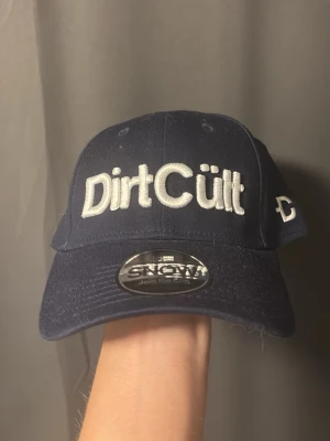 Mörkblå DirtCült keps - Snygg mörkblå keps från DirtCült med broderad vit logga framtill och justerbar rem bak. Klassisk böjd skärm och sexpanelers design. Tillverkad i bomullstyg som ger en skön och avslappnad look.