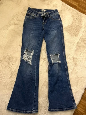 Blå lågmidjade bootcut jeans med slitningar 157 - Snygga blå bootcut jeans från 157 med slitna hål på båda knäna. Jeansen har klassisk femficksdesign, normalhög midja och utställda ben. Perfekta för dig som gillar en avslappnad och trendig stil. Lågmidjade 