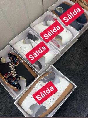 Svarta sneakers med färgstänk - Unika svarta sneakers från Maison Margiela med färgstänk i rött, blått, gult och vitt. Skorna har vita snören, brun sula och en klassisk låg siluett. Perfekta för dig som vill sticka ut med din stil och gillar konstnärliga detaljer.