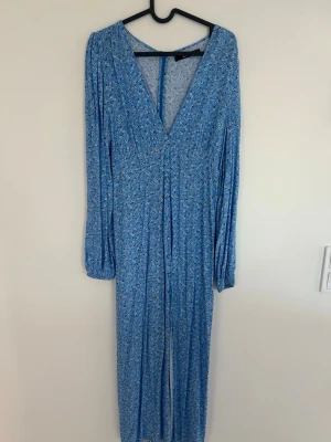 Blommig blå långärmad klänning  - Supersöt blå klänning med små vita blommor, v-ringning och långa ärmar med resår vid handlederna. Dragkedja i ryggen 