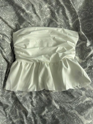 Vit bandeau tubtopp med volang - Säljer en vit bandeau tubtopp med volang nertill. Toppen är axelbandslös och har en snygg rynkad design upptill som ger en cool och trendig look. Perfekt för dig som vill ha något stilrent och enkelt att matcha.