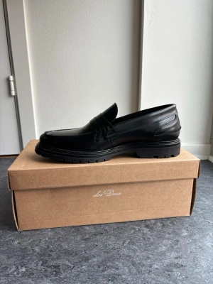 Svarta loafers från Les Deux - Stilrena svarta loafers från Les Deux i slätt läder med chunky sula och klassisk penny-detalj över vristen. Skorna har en modern siluett och grov yttersula som ger ett trendigt intryck. Perfekta för dig som vill ha en clean look med edge.