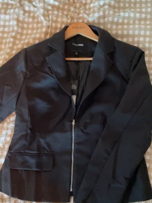 Svart kavaj med dragkedja Fashion Moda - Stilren svart kavaj från Fashion Nova med dragkedja framtill istället för knappar. Klassisk krage, figurnära passform och två fickor framtill. 