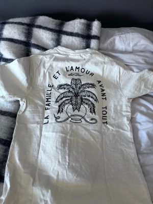 Vit t-shirt från Les Deux med tryck - Säljer en vit t-shirt från Les Deux med svart tryck på ryggen med texten 'La famille et l'amour avant tout' och palm-motiv. Liten logga på bröstet. Klassisk passform och rund hals. Perfekt till jeans eller shorts.