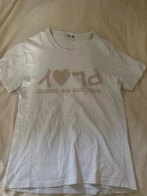 Vit Comme des Garçons PLAY t-shirt - Säljer en vit t-shirt från Comme des Garçons PLAY med beige tryck framtill. Klassisk rund hals och korta ärmar. T-shirten har det ikoniska PLAY-trycket med hjärta och text. Perfekt för dig som gillar stilrena och trendiga plagg.