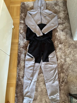 Under Armour grå och svart loungewear - Säljer ett set från Under Armour med hoodie och byxor i grått och svart. Hoodien har dragkedja och fickor, byxorna har resår i midjan och en praktisk ficka med dragkedja. Perfekt för chill dagar hemma eller när du vill vara bekväm.