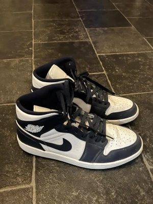 Nike Air Jordan 1 Mid svart/vit - Säljer ett par Nike Air Jordan 1 Mid sneakers i svart och vitt. Klassisk design med hög siluett, perforerad tå och ikonisk Swoosh på sidan. Skorna har svarta skosnören och är tillverkade i läder och textil. Perfekt för dig som vill ha en tidlös och cool look.