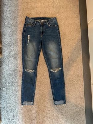 Blå jeans - Väldigt fina jeans med hål för knäna. Använda men har mycket kvar att ge. Väldigt bekväma! Lite speciella vid anklarna då de är längre bak än fram. Raka i modellen.