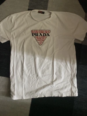Vit t-shirt från Prada med tryck - Säljer en vit t-shirt från Prada med ett stort trekantigt tryck på bröstet där det står PRADA och text i rött och svart. Klassisk rund hals och korta ärmar. Perfekt för dig som gillar stilrena plagg med exklusiv känsla.