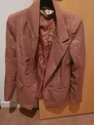 Kavaj - Snygg brun kavaj med dubbelknäppning och breda slag. Kavajen har två framfickor med lock och är helfodrad. Passar perfekt till en trendig och stilren look. Materialet ser ut att vara polyester eller liknande tyg. Det är i kinesiskt storlek men motsvarar svensk storlek 34/36