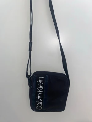 Svart axelväska från Calvin Klein - Snygg svart axelväska från Calvin Klein med justerbar rem och vit logga på framsidan. Väskan har en fyrkantig form och är tillverkad i slitstarkt tygmaterial. Perfekt för att bära det viktigaste och ger en clean streetstyle-look..