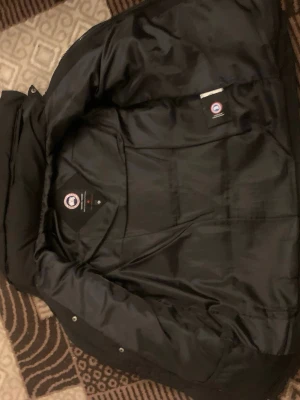 Svart dunjacka från Canada Goose - Säljer en svart dunjacka från Canada Goose med klassisk logotyp på ärmen. Jackan har en hög krage, avtagbar huva och knäppning med både dragkedja och tryckknappar. Perfekt för kalla vinterdagar och riktigt varm tack vare dunfyllningen.