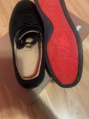 Svarta Louboutin sneakers med röd sula - Snygga svarta sneakers från Christian Louboutin med klassisk röd sula och svart mocka. Skorna har svarta skosnören, låg profil och beige innersula med logga. Perfekta för dig som vill sticka ut med exklusiv stil.